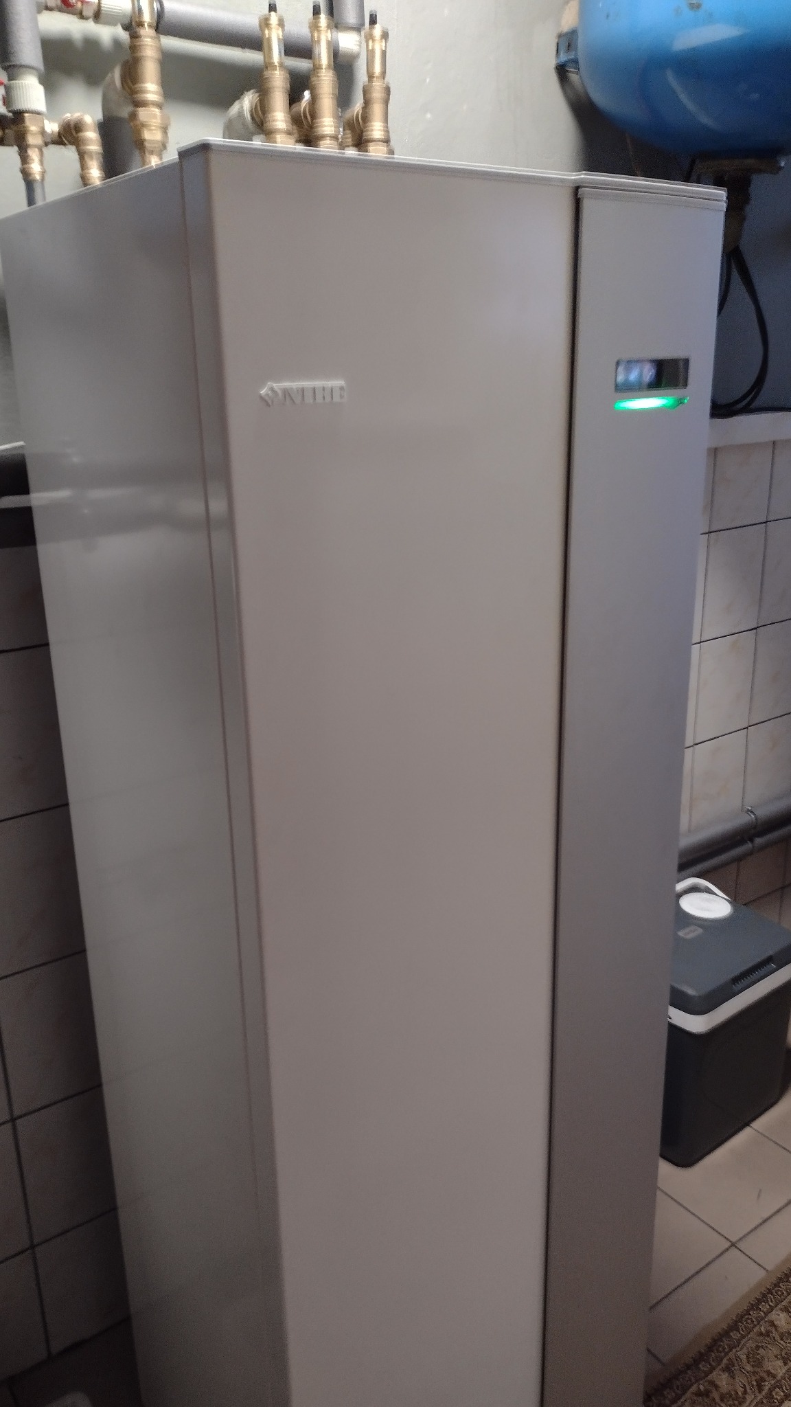 Biała jednostka wewnętrzna z widocznym logo Nibe i zieloną diodą, z instalacją hydrauliczną na górze, stojąca w pomieszczeniu wyłożonym kafelkami.