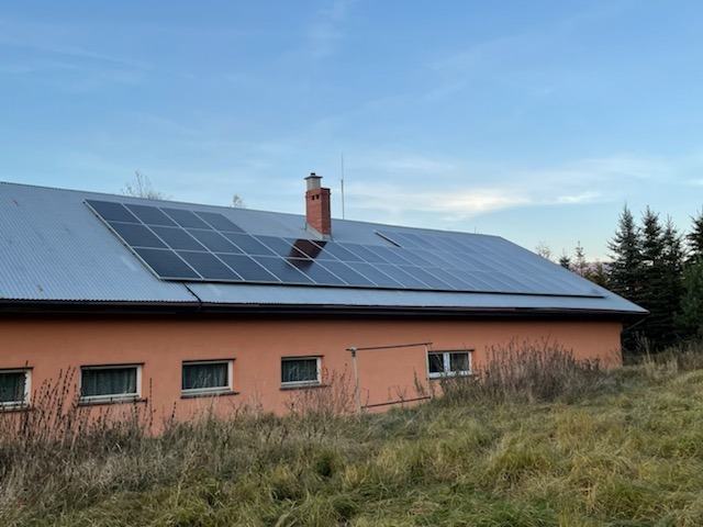 Instalacja fotowoltaiczna na dachu budynku z blachy falistej, widok z dołu na tle pochmurnego nieba i drzew iglastych.