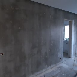 jacek nowak - Surowa ściana po nałożeniu gładzi, widoczne puszki elektryczne, w tle pracownik w kombinezonie malarskim i drabina, pomieszczenie w trakcie prac wykończeniowych.