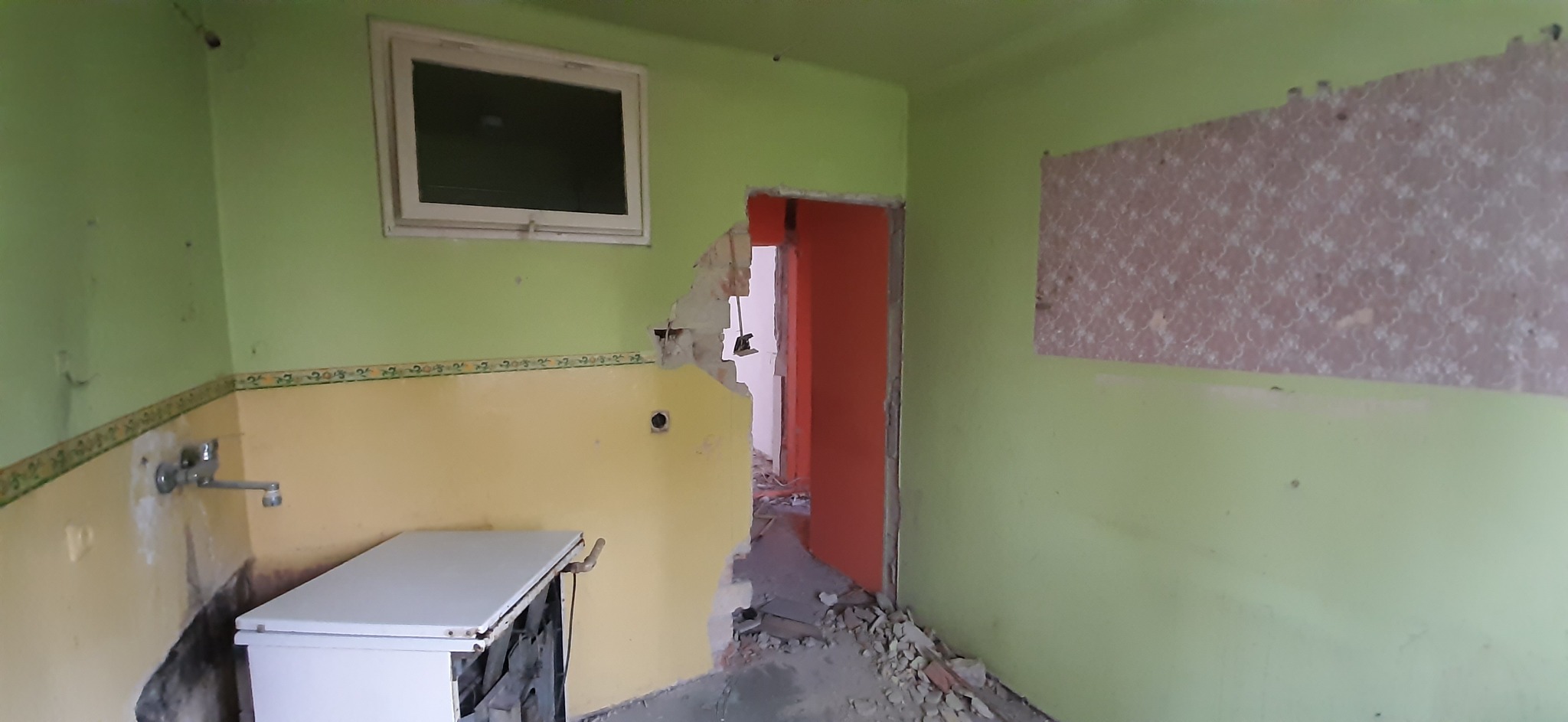 Kuchnia w trakcie demontażu: ściany w odcieniach zieleni i żółci z fragmentami zerwanych płytek, widoczny zdemontowany fragment ściany przy wejściu do innego pomieszczenia z pomarańczowymi drzwiami...