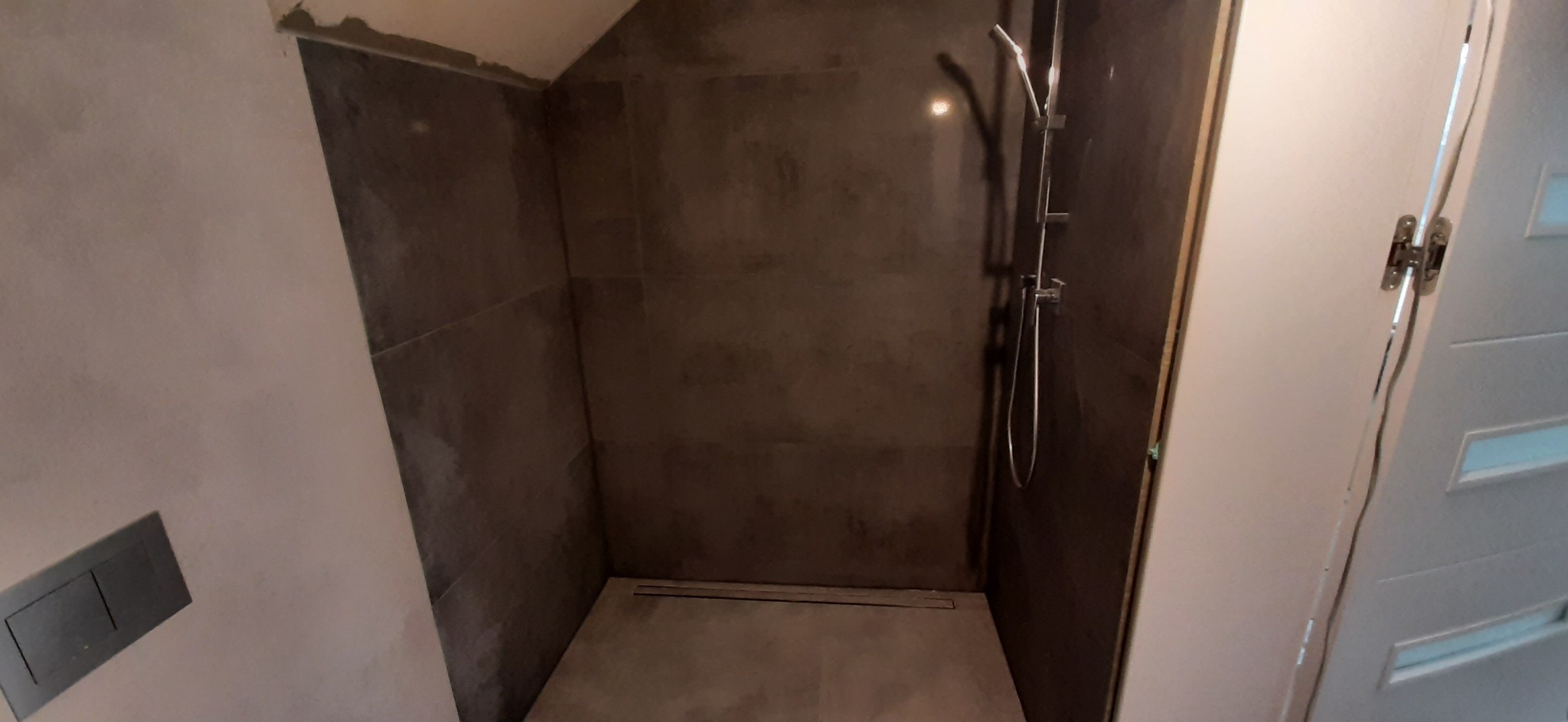 Nowoczesna kabina prysznicowa z szarymi płytkami i odpływem liniowym, widok z perspektywy drzwi z zamontowanymi zawiasami.