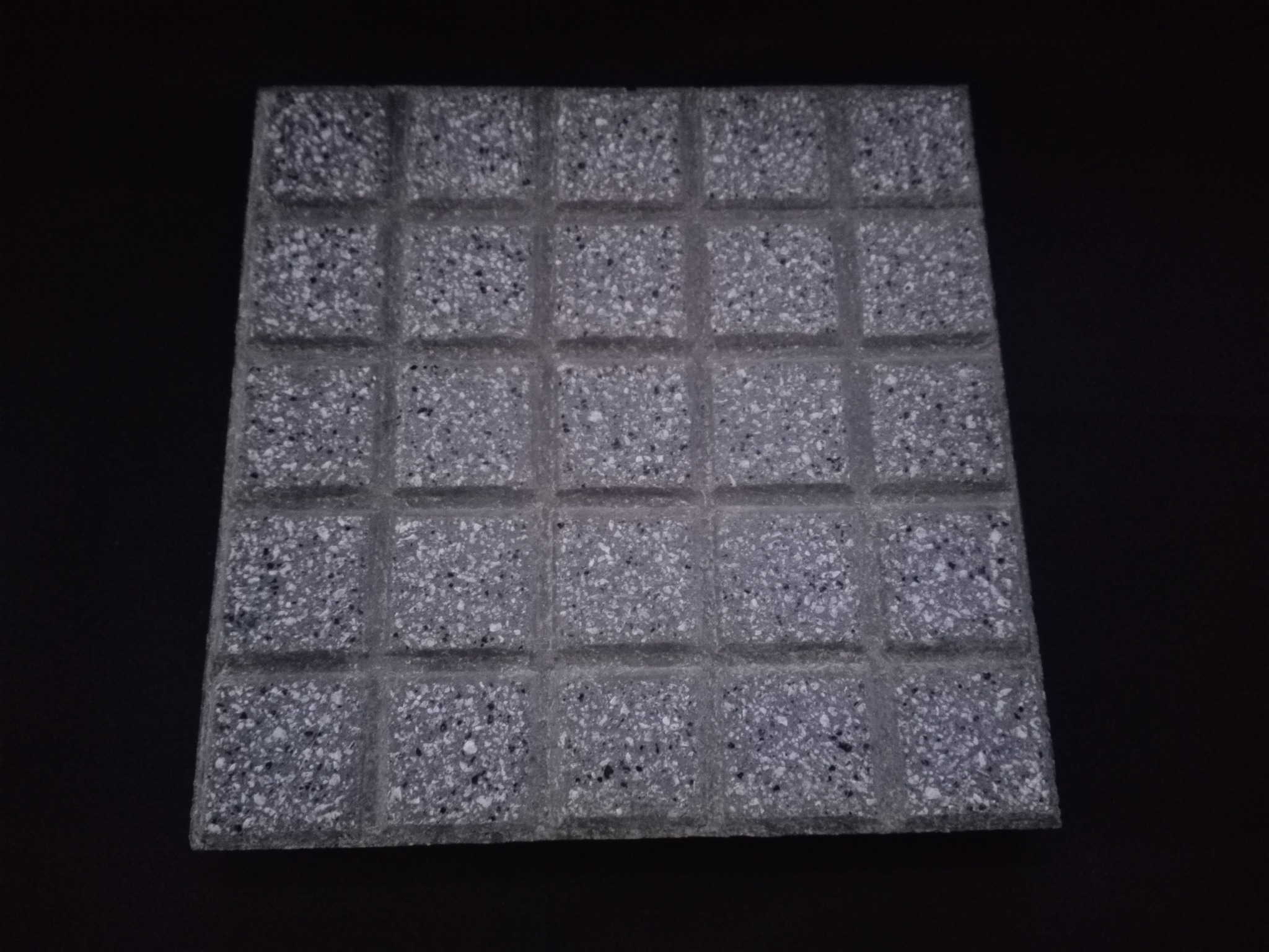 Kwadratowa, szara płyta chodnikowa z drobnymi, ciemnymi i jasnymi kamyczkami, podzielona na 36 mniejszych kwadratów, leży na czarnym tle.