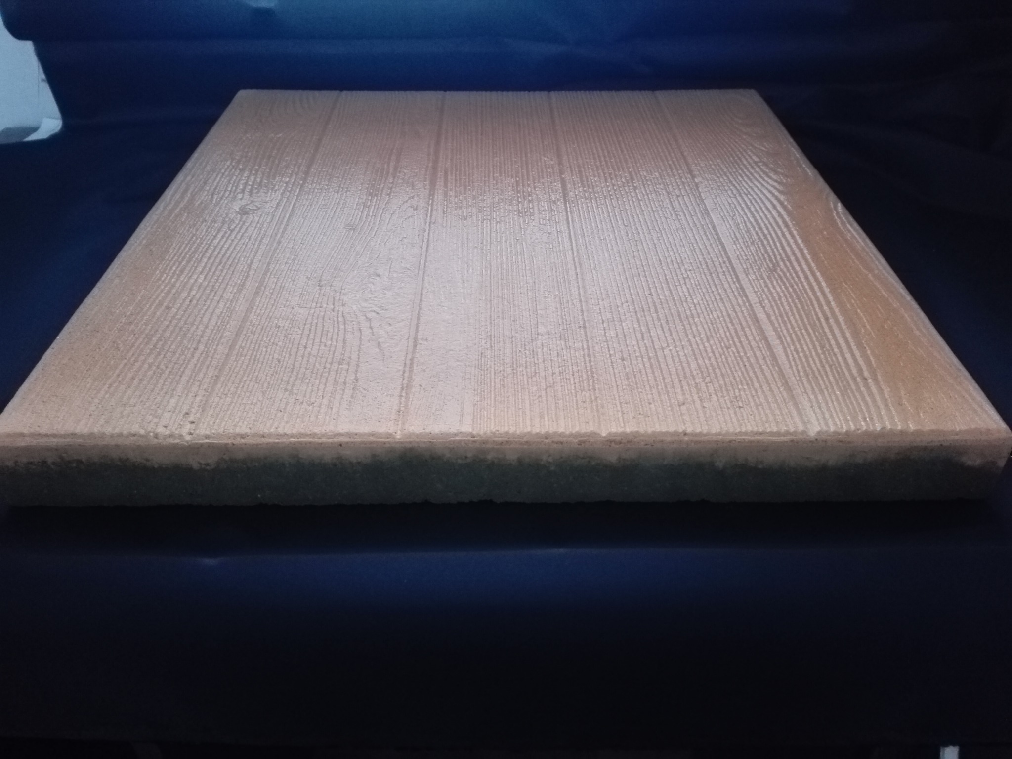 Beżowa płyta chodnikowa z imitacją drewna na ciemnym tle, widoczna struktura i krawędzie.