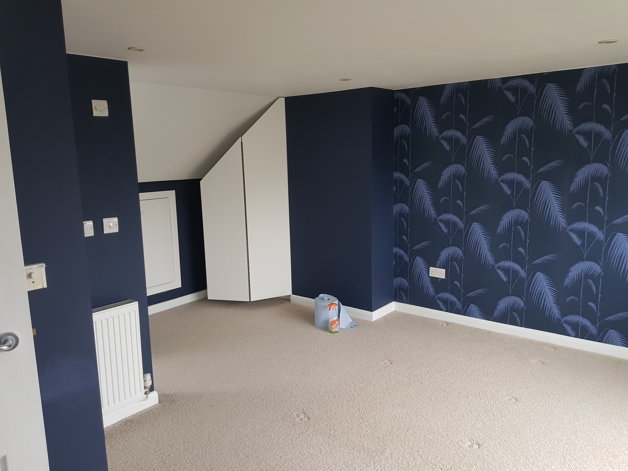 Pokój po odświeżeniu: ściany w kolorze granatowym, jedna z nich z tapetą w motyw liści palmowych, jasna wykładzina dywanowa, białe szafy wnękowe pod skosem.