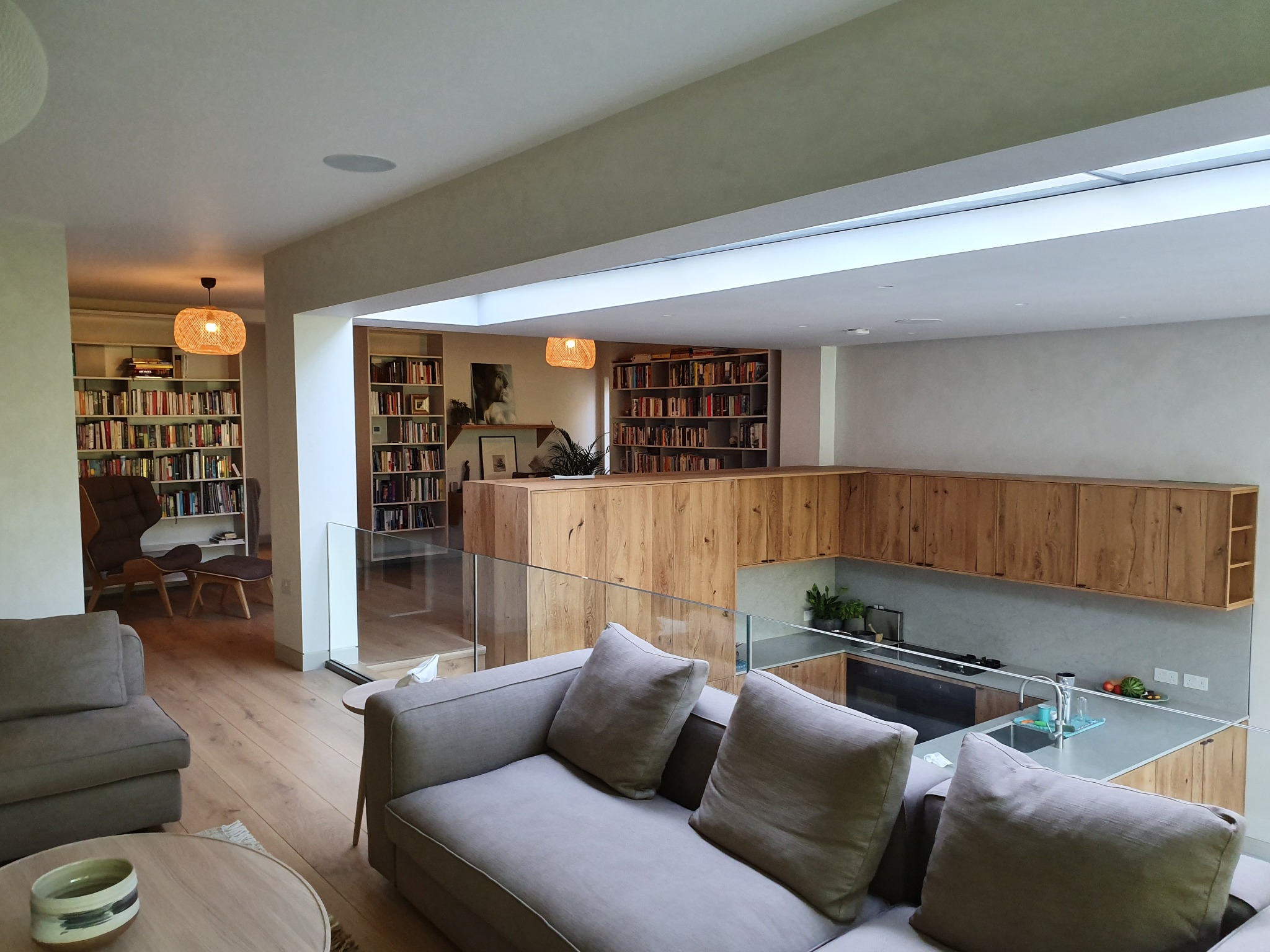 Przestronne wnętrze domu z antresolą, biblioteczkami i drewnianą zabudową kuchenną, oświetlone naturalnym światłem z okna dachowego.