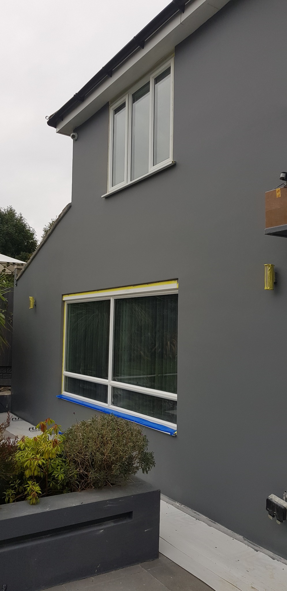 Świeżo pomalowana szara elewacja domu z białymi oknami, zabezpieczonymi taśmą malarską, widoczne detale ogrodu.