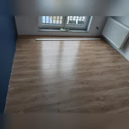 Jasne panele podłogowe z widoczną strukturą drewna, ułożone w pomieszczeniu z niebieską ścianą i białym grzejnikiem pod oknem, listwa przypodłogowa jeszcze nie zamontowana.