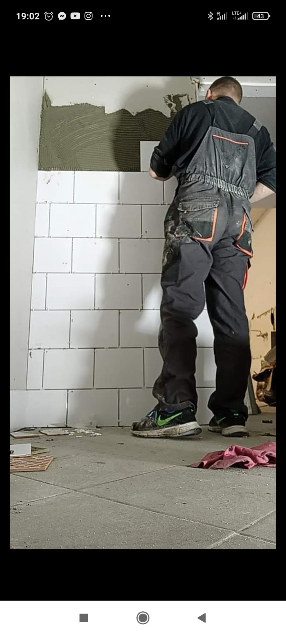 Mężczyzna w roboczym kombinezonie układa białe, prostokątne płytki na ścianie pokrytej klejem, widoczne brudne buty sportowe i fragmenty płytek na podłodze.