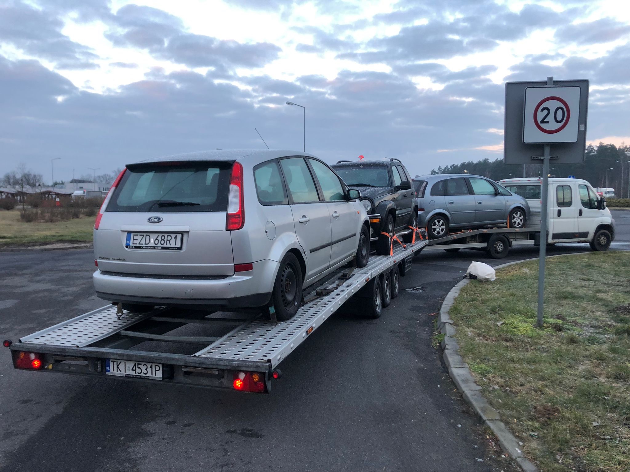 Laweta z trzema samochodami osobowymi, w tym srebrny Ford Focus z tyłu, na tle szarego, pochmurnego nieba i znaku drogowego z ograniczeniem prędkości do 20 km/h.