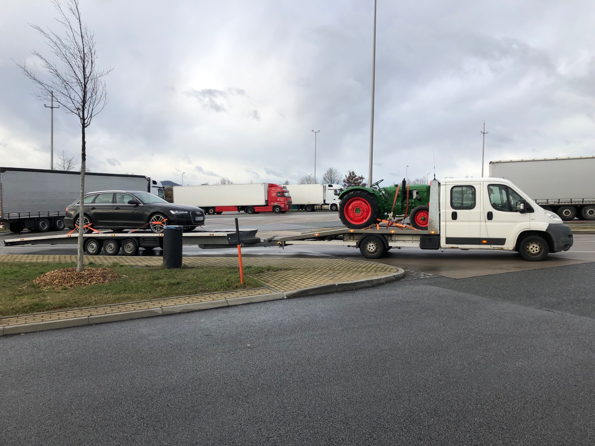 Laweta z białym busem przewożąca czarne Audi kombi i zielony traktor z czerwonymi kołami na parkingu dla tirów, pochmurne niebo w tle.