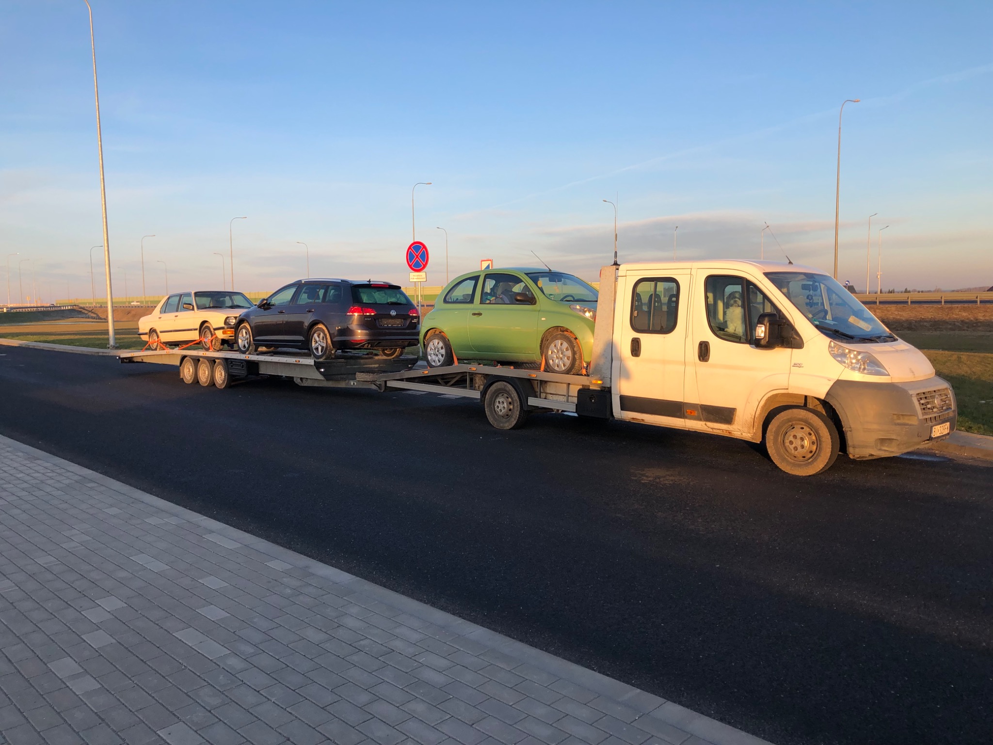 Laweta z trzema samochodami różnych marek i kolorów: beżowe BMW, szary Volkswagen Golf i zielony Nissan Micra, zaparkowana na poboczu drogi z widocznym znakiem zakazu parkowania.