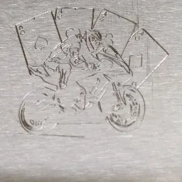 Grawerowany motocyklista na metalu otoczony asami kart, wykonany prawdopodobnie laserem.