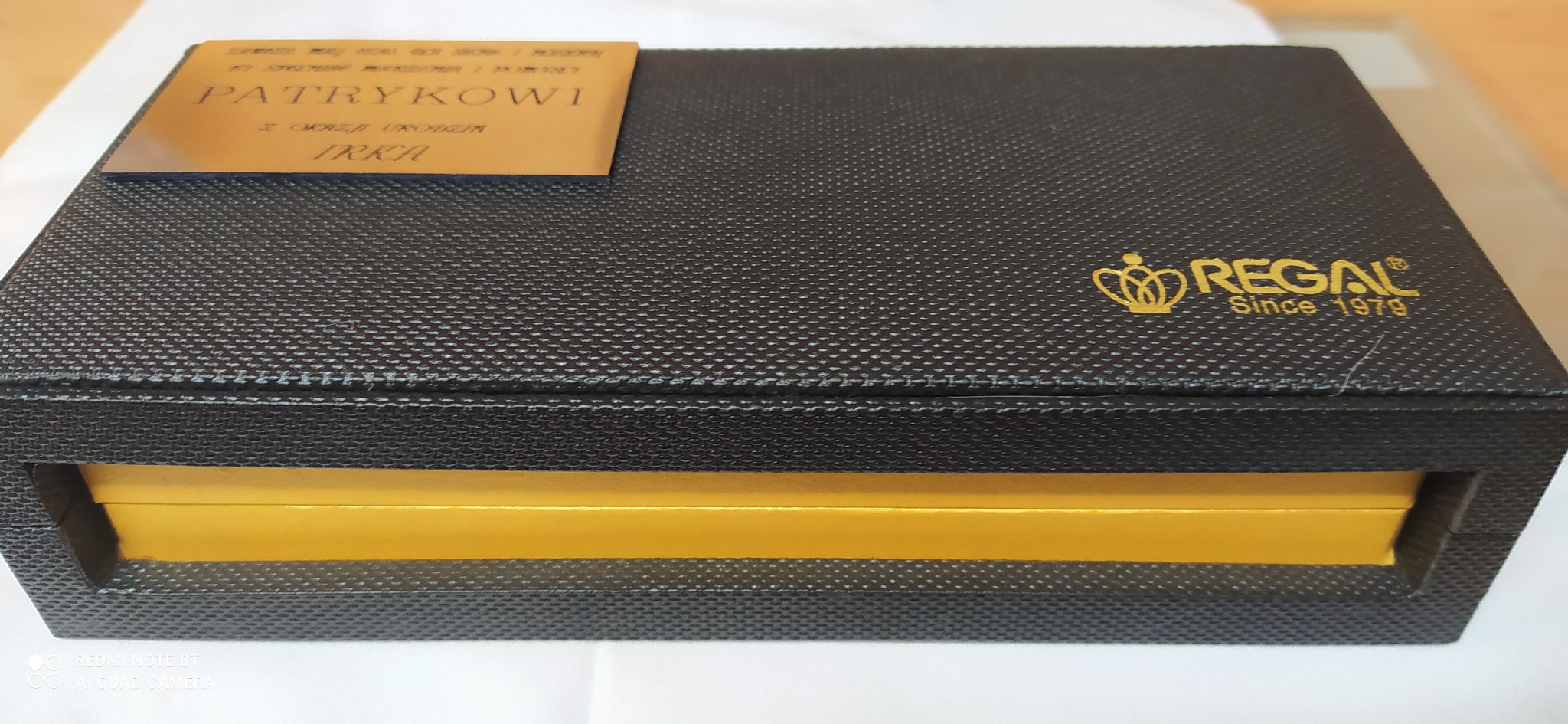 Czarno-złote pudełko prezentowe z teksturowaną powierzchnią, spersonalizowane złotą tabliczką z napisem 'Patrykowi z okazji urodzin Irka', z logo firmy 'Regal Since 1979' w prawym górnym rogu.