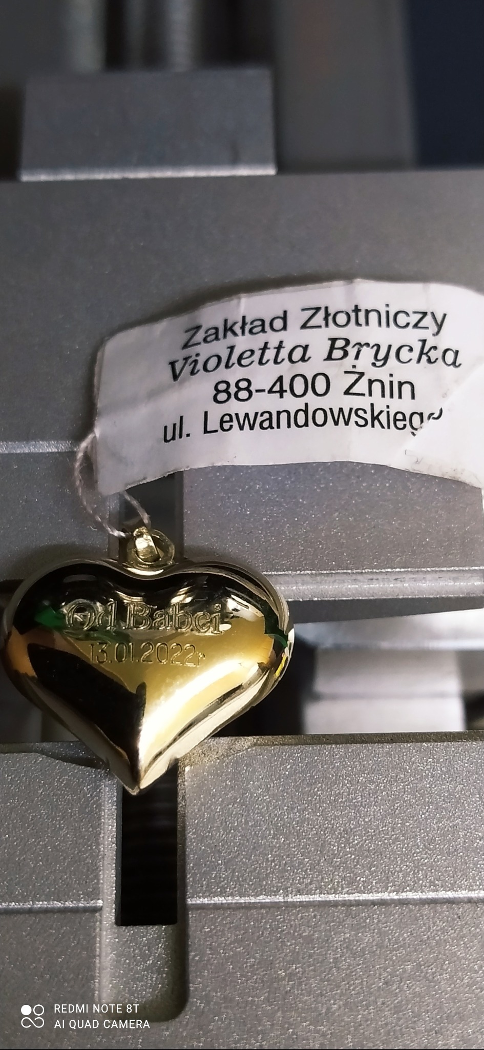 Złoty wisiorek w kształcie serca z wygrawerowanym napisem 'Od Babci 13.01.2022' na tle etykiety Zakładu Złotniczego Violetta Brycka z Żnina, prezentowany na metalowej powierzchni.