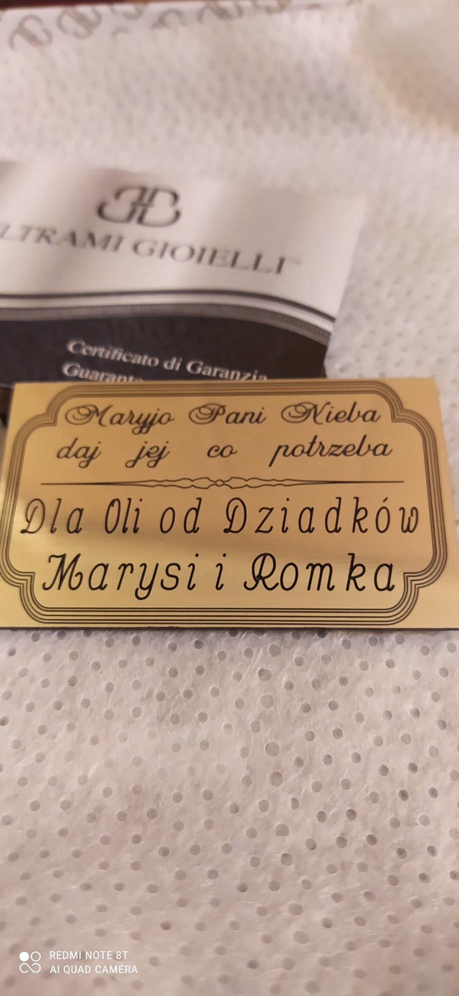 Złota tabliczka z dedykacją 'Maryjo Pani Nieba daj jej co potrzeba Dla Oli od Dziadków Marysi i Romka' na tle certyfikatu firmy Ultrami Gioielli.