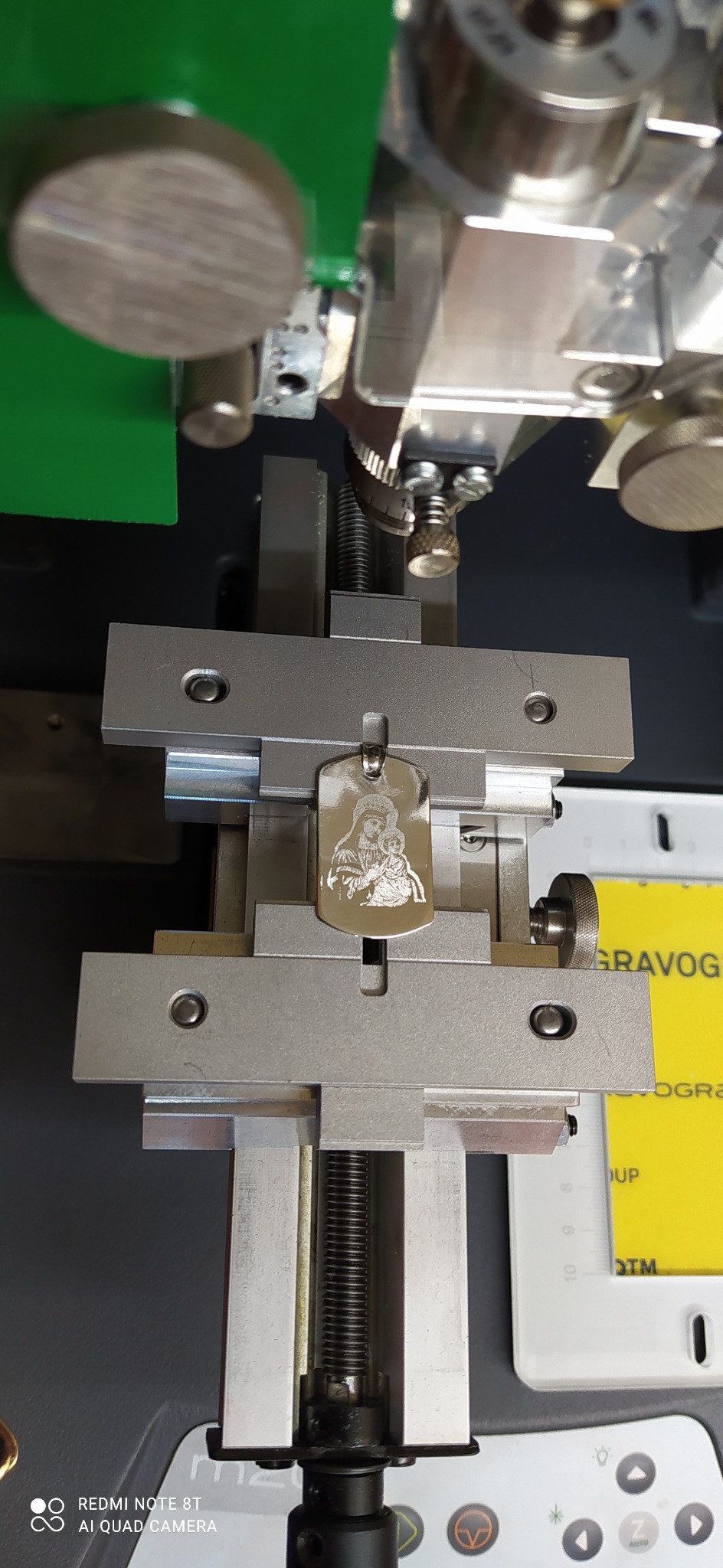 Metalowy brelok z wygrawerowanym wizerunkiem Matki Boskiej z Dzieciątkiem zamocowany w maszynie grawerującej.