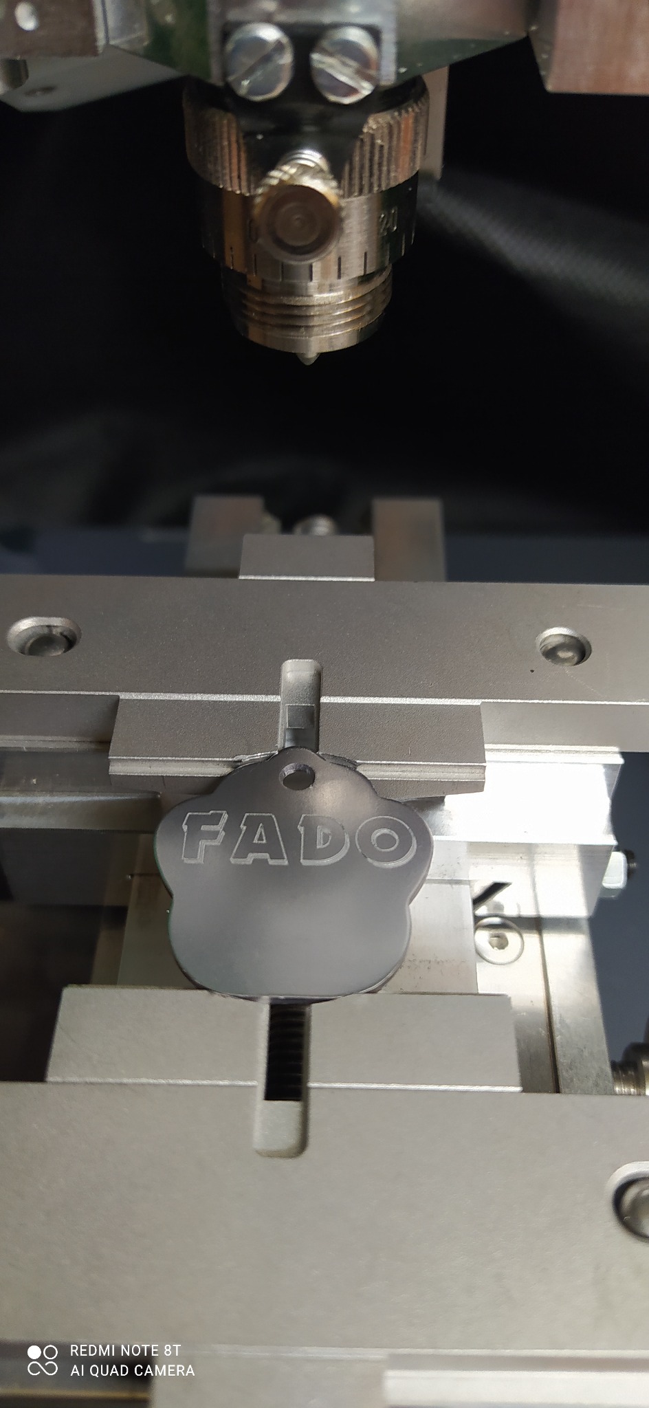 Proces grawerowania laserowego napisu 'FADO' na metalowym breloczku o nieregularnym kształcie, umieszczonym w precyzyjnym uchwycie maszyny.