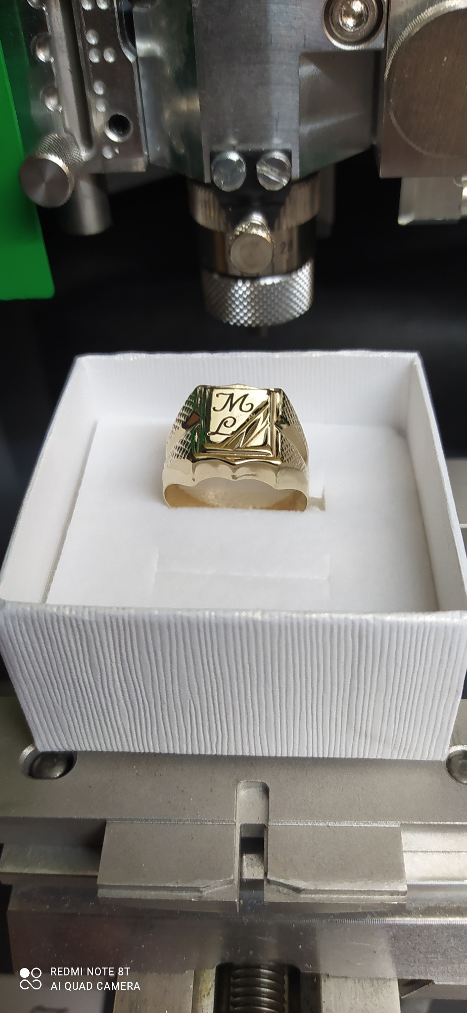 Złoty sygnet z wygrawerowanymi inicjałami 'ML' w białym pudełku, umieszczony wewnątrz maszyny do grawerowania laserowego.
