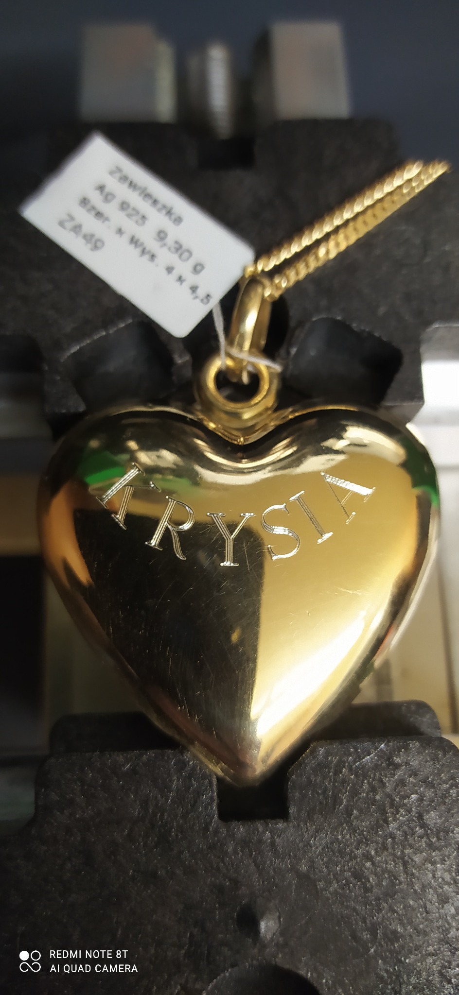Złota zawieszka w kształcie serca z wygrawerowanym imieniem 'Krysia', zawieszona na łańcuszku, z dołączoną metką z opisem kruszcu i wagi.