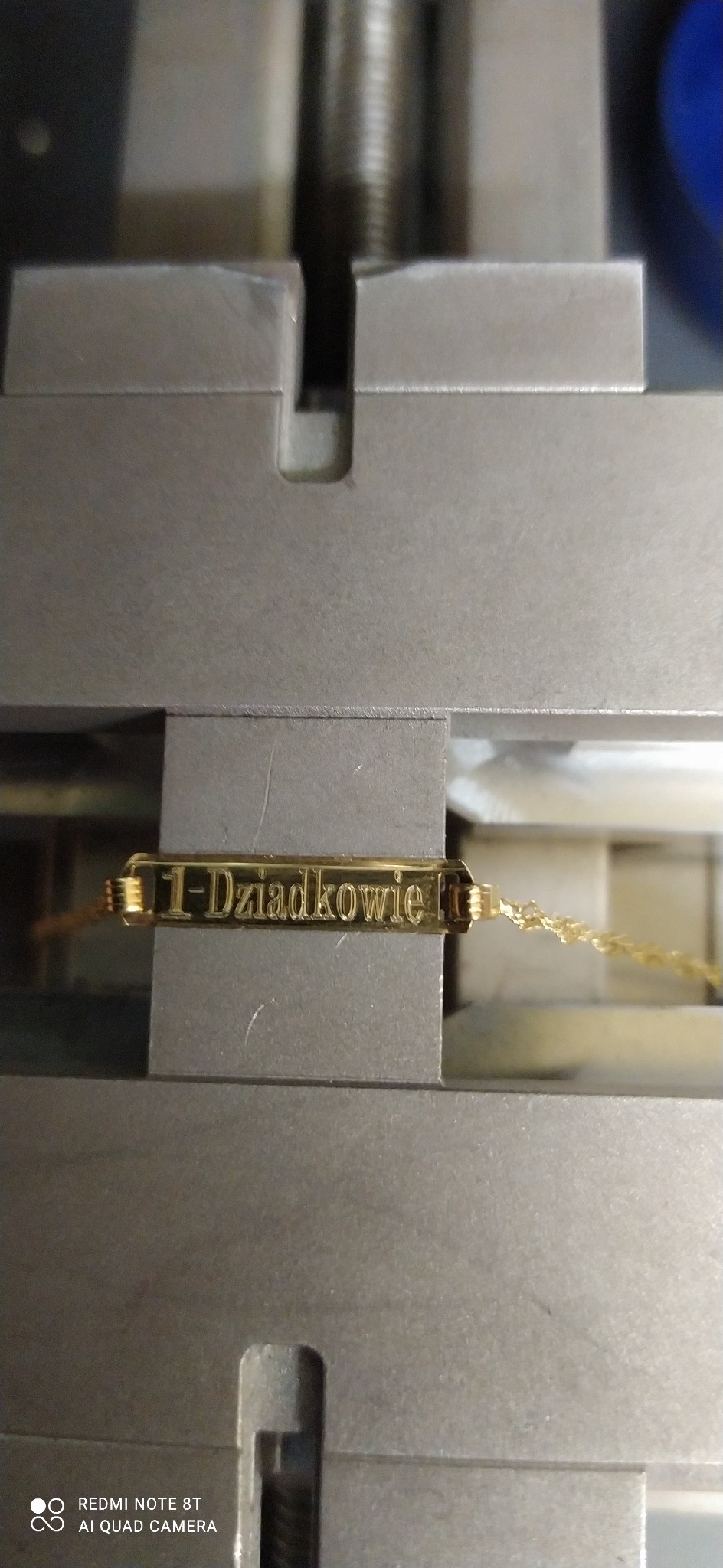 Złota bransoletka z napisem '1-Dziadkowie' umieszczona w metalowym uchwycie maszyny grawerującej, zbliżenie na grawerowany napis.
