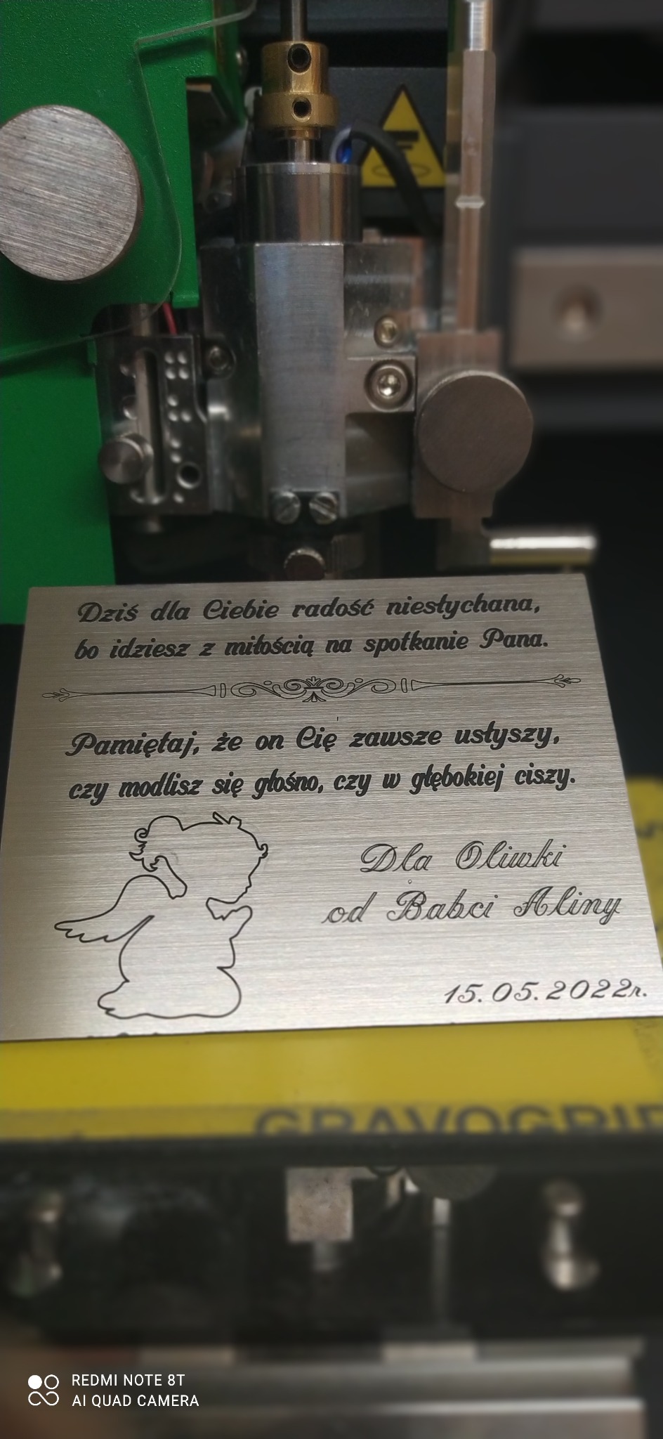 Metalowa tabliczka z wygrawerowanym tekstem dedykacji i rysunkiem anioła, umieszczona w maszynie grawerującej. Widoczny fragment mechanizmu urządzenia.