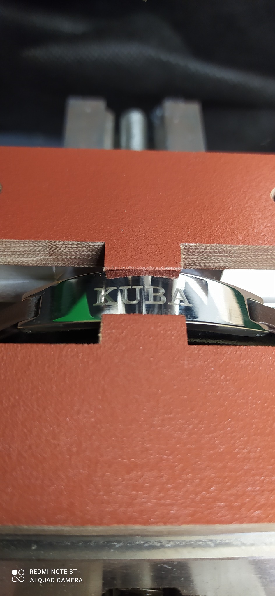 Metalowy element z wygrawerowanym napisem 'KUBA' zamocowany w imadle z brązowymi szczękami.