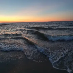 Spokojne fale morskie o zachodzie słońca, delikatne refleksy pomarańczowego światła na wodzie, widoczne drewniane pale w oddali.