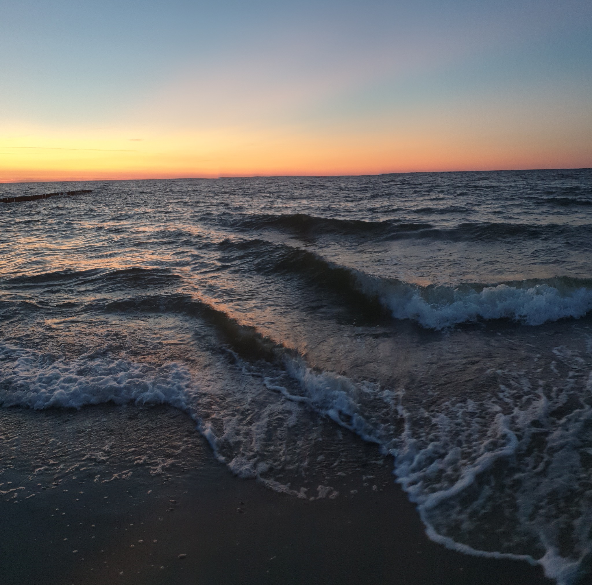 Spokojne fale morskie o zachodzie słońca, delikatne refleksy pomarańczowego światła na wodzie, widoczne drewniane pale w oddali.