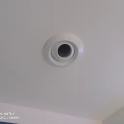 Biały, okrągły nawiewnik sufitowy z regulowaną dyszą, zamontowany w białym suficie. Widoczny fragment niebieskiej ściany w prawym dolnym rogu.