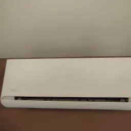 Zamontowany, biały klimatyzator Midea na brązowej ścianie, widoczny z bliska. Górna część ściany kremowa. Widoczne filtry i kratka wlotu powietrza.