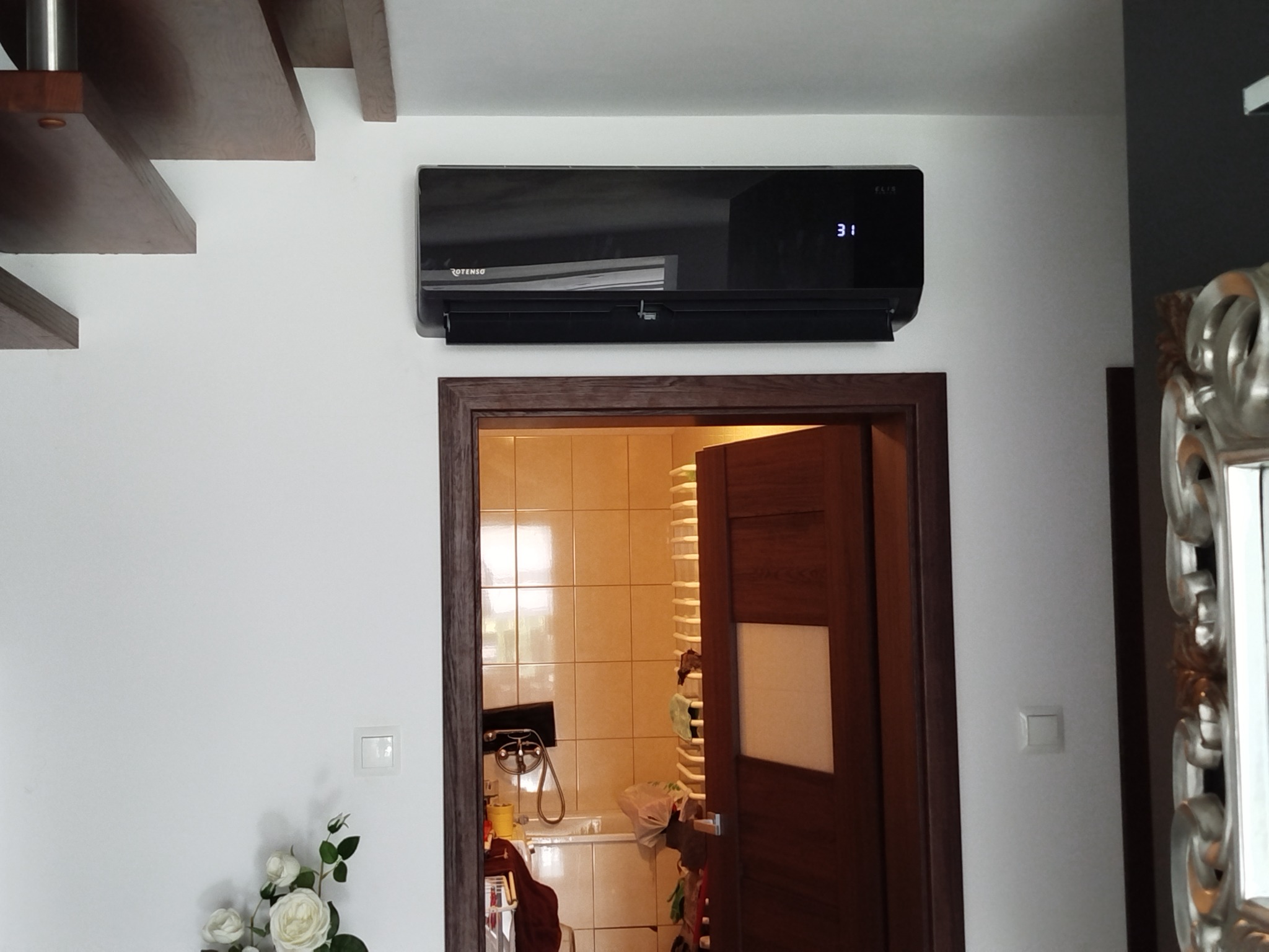 Czarny klimatyzator Rotenso na białej ścianie, nad brązowymi drzwiami do łazienki. Widoczne schody i dekoracja kwiatowa. Ustawiona temperatura 31 stopni.
