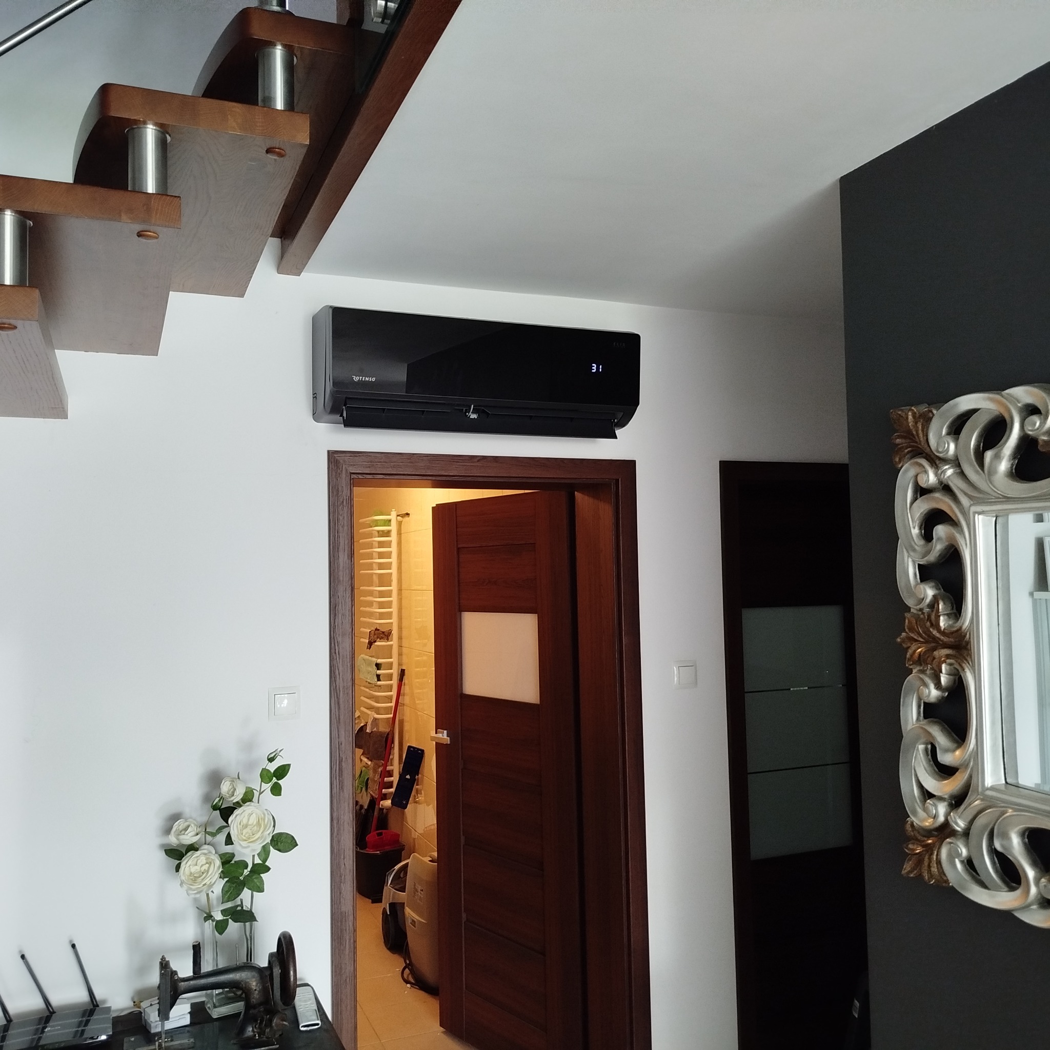 Czarny klimatyzator Rotenso na białej ścianie w nowoczesnym wnętrzu, widok z dołu. W tle schody z drewnianymi stopniami i lustro w ozdobnej ramie.