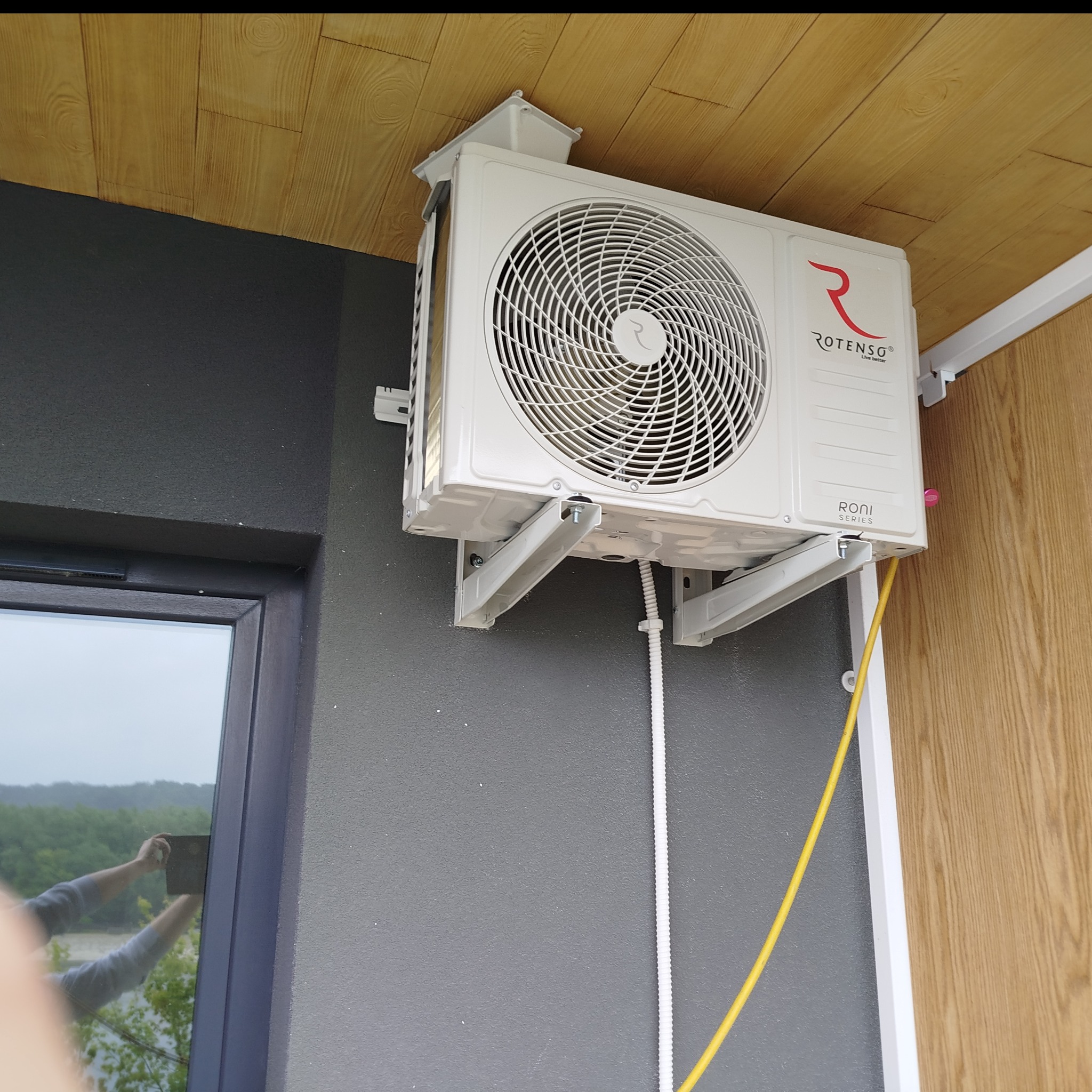 Zamontowana klimatyzacja Rotenso Roni Series na szarej ścianie zewnętrznej budynku, widoczne przewody i uchwyty montażowe, odbicie fotografa w oknie.