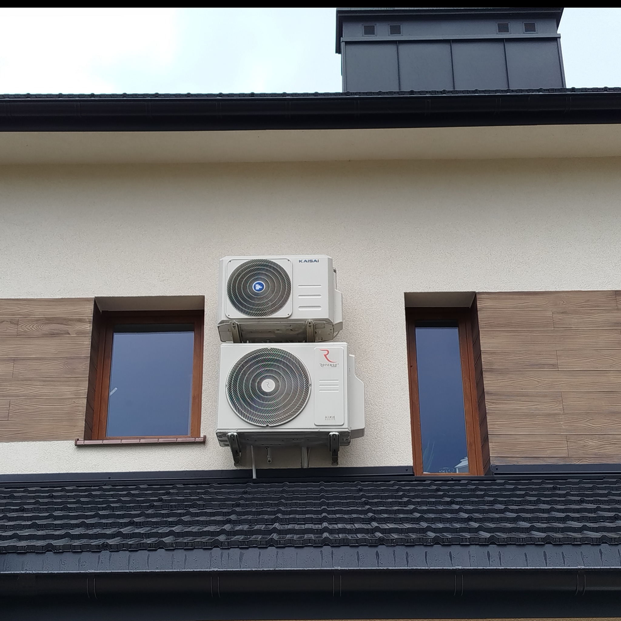 Dwa klimatyzatory marki Kaisai i Rotenso zamontowane na elewacji budynku z oknami w drewnianych ramach i ciemnym dachem. Widok z dołu.