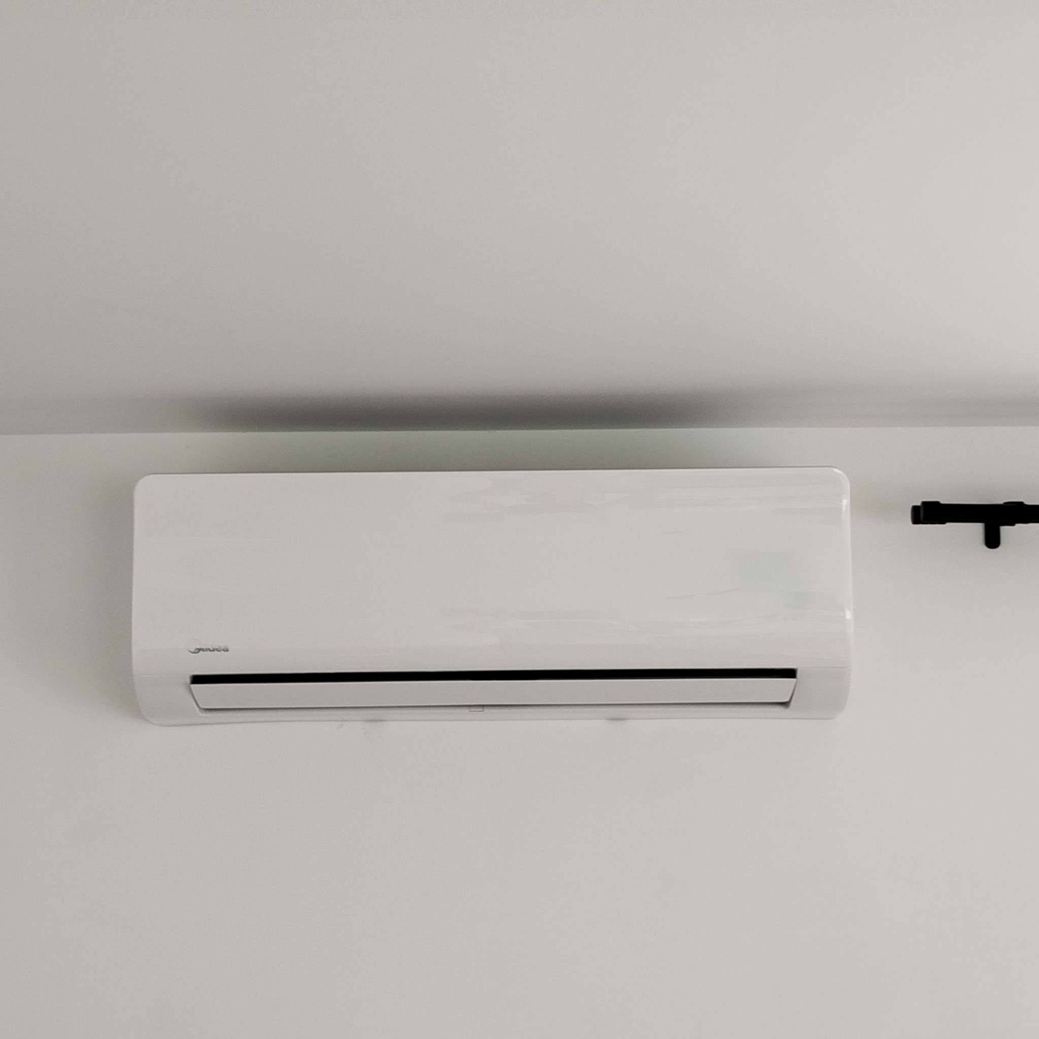 Biała klimatyzacja Midea na jasnej ścianie, widoczny fragment czarnej rury. Minimalistyczny design, stonowane oświetlenie.