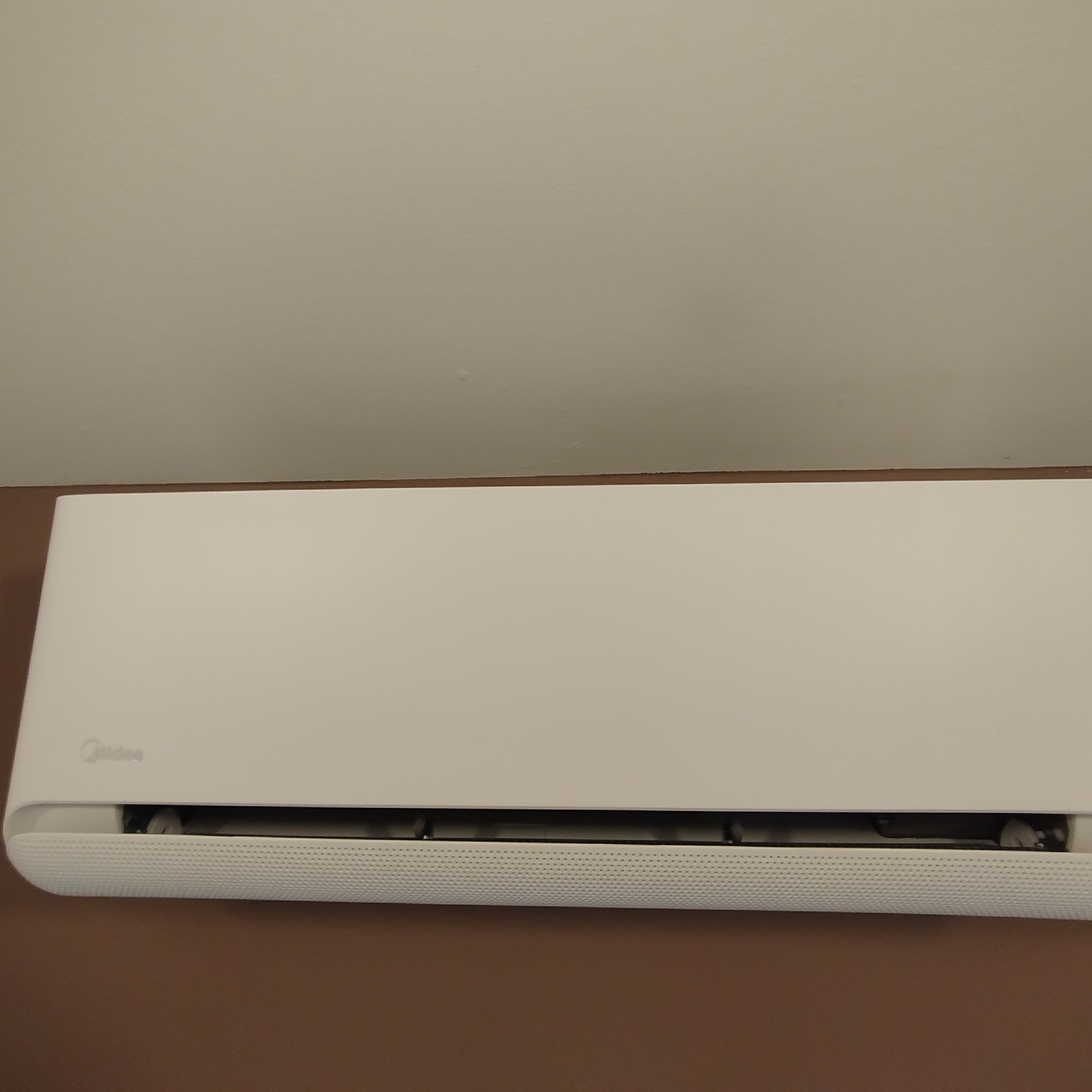 Zamontowany, biały klimatyzator Midea na brązowej ścianie, widoczny z bliska. Górna część ściany kremowa. Widoczne filtry i kratka wlotu powietrza.