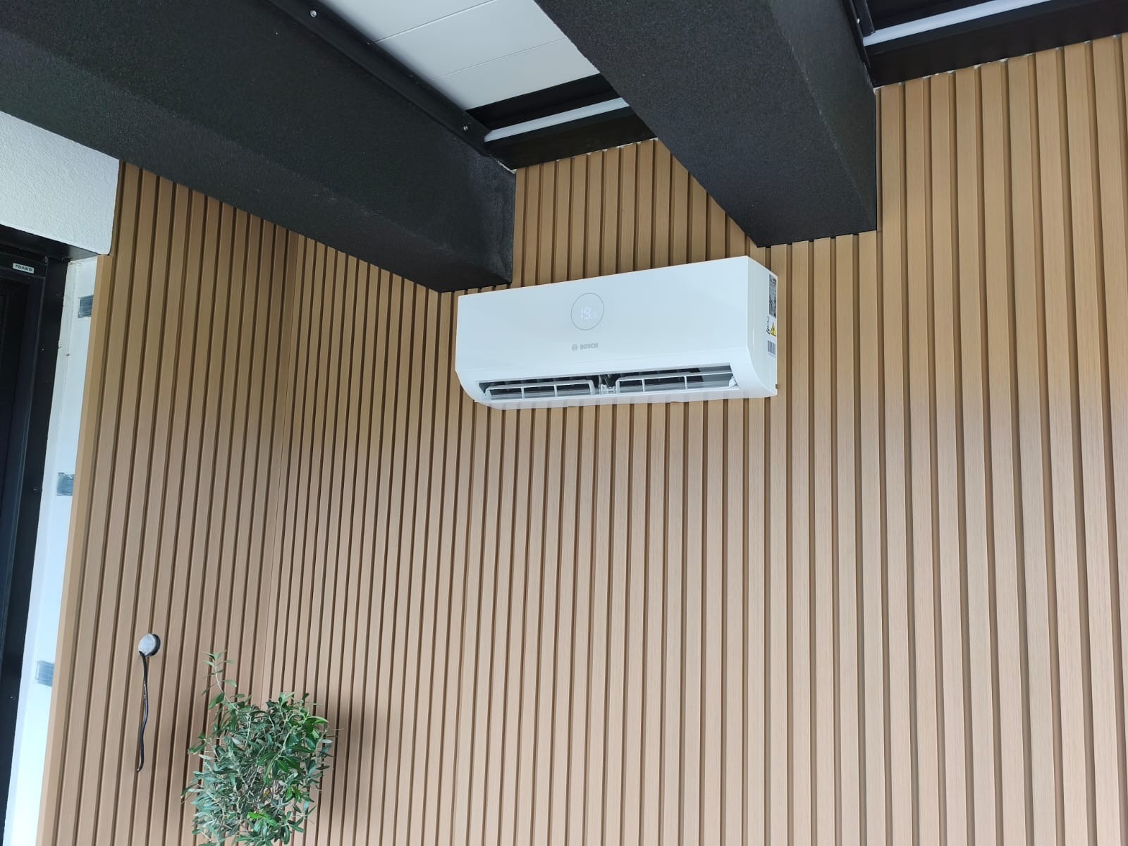 Biała klimatyzacja Bosch zamontowana na ścianie z drewnianych paneli, widoczna zieleń rośliny doniczkowej i czarne elementy sufitu. Nowoczesny design.