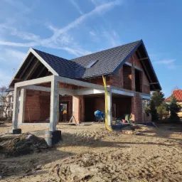 Budowa domu jednorodzinnego w stanie surowym otwartym: ściany z czerwonej cegły, betonowe słupy pod zadaszeniem, czarny dach z oknem dachowym, niebieska betoniarka i piasek na placu budowy.