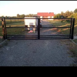 Nowoczesna, czarna, metalowa brama wjazdowa z siatki, prowadząca na posesję z domem z czerwoną dachówką i zaparkowaną przyczepą.