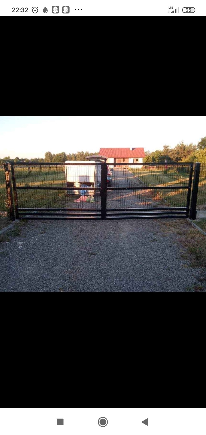 Nowoczesna, czarna, metalowa brama wjazdowa z siatki, prowadząca na posesję z domem z czerwoną dachówką i zaparkowaną przyczepą.