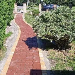 Czerwona ścieżka z kostki brukowej z żółtymi obrzeżami, wijąca się między zielonymi krzewami i trawnikiem w słoneczny dzień.