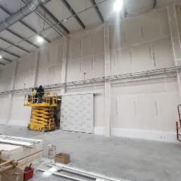 Montaż płyt warstwowych wewnątrz hali przemysłowej: pracownicy na podnośniku nożycowym mocują płyty do ściany, widoczne elementy konstrukcji stalowej i instalacje pod sufitem.