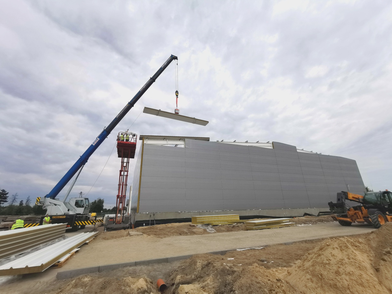 Montaż płyt warstwowych na dachu hali magazynowej przy użyciu dźwigu i podnośnika koszowego, widoczne ułożone płyty warstwowe i maszyny budowlane na placu budowy.