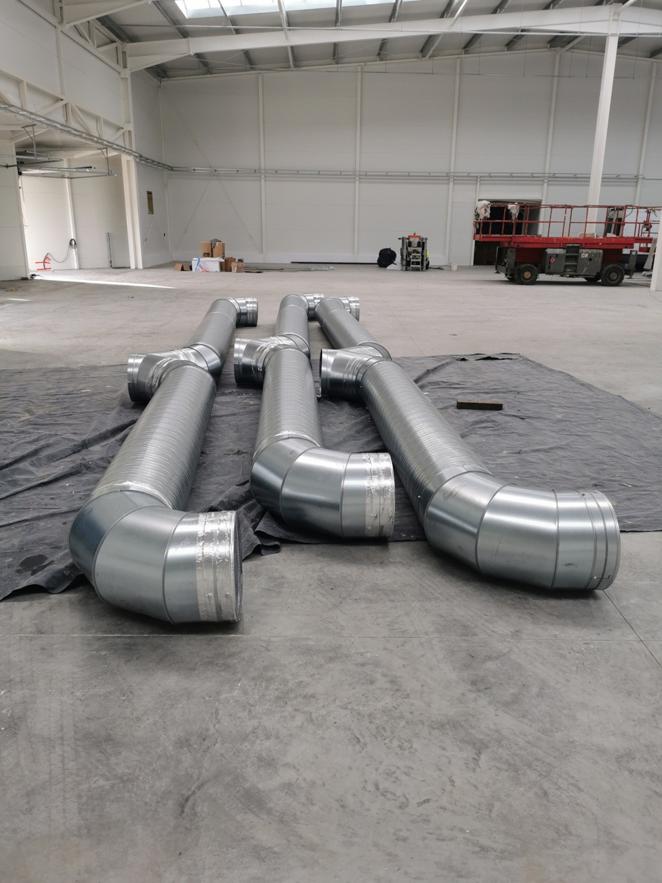 Elementy układu wentylacyjnego z blachy ocynkowanej, przygotowane do montażu w hali magazynowej z widocznym podnośnikiem koszowym w tle.