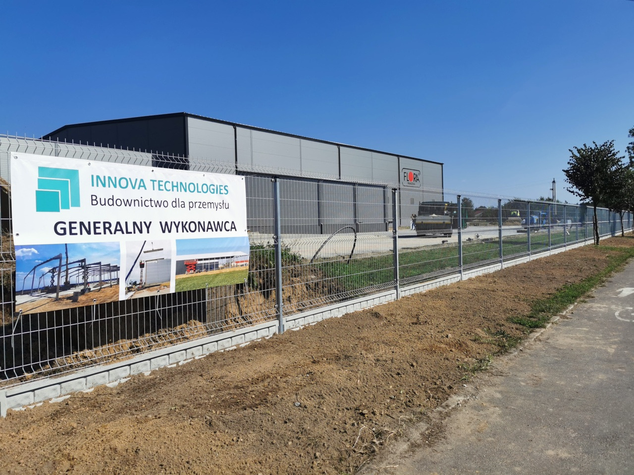 Hala przemysłowa z szarej blachy trapezowej, widoczna zza ogrodzenia z siatki, na którym umieszczono baner firmy Innova Technologies, generalnego wykonawcy budownictwa dla przemysłu, z miniaturami...