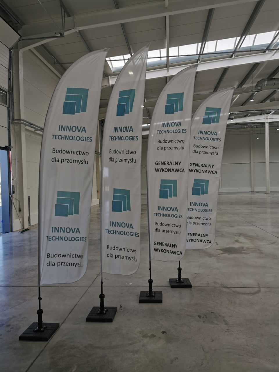 Cztery identyczne, białe, łukowate flagi reklamowe z logo i napisem 'INNOVA TECHNOLOGIES Budownictwo dla przemysłu' oraz 'GENERALNY WYKONAWCA' ustawione rzędem w hali przemysłowej.