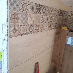 Ściana łazienki wyłożona beżowymi płytkami z dekoracyjnym pasem płytek wzorzystych, widoczny poziomica i fragment toalety.