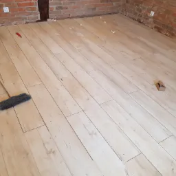 Ułożone jasne panele podłogowe imitujące drewno w pomieszczeniu z fragmentami odsłoniętej cegły na ścianach, widoczne narzędzie do cięcia, szczotka do zamiatania i resztki po cięciu paneli.