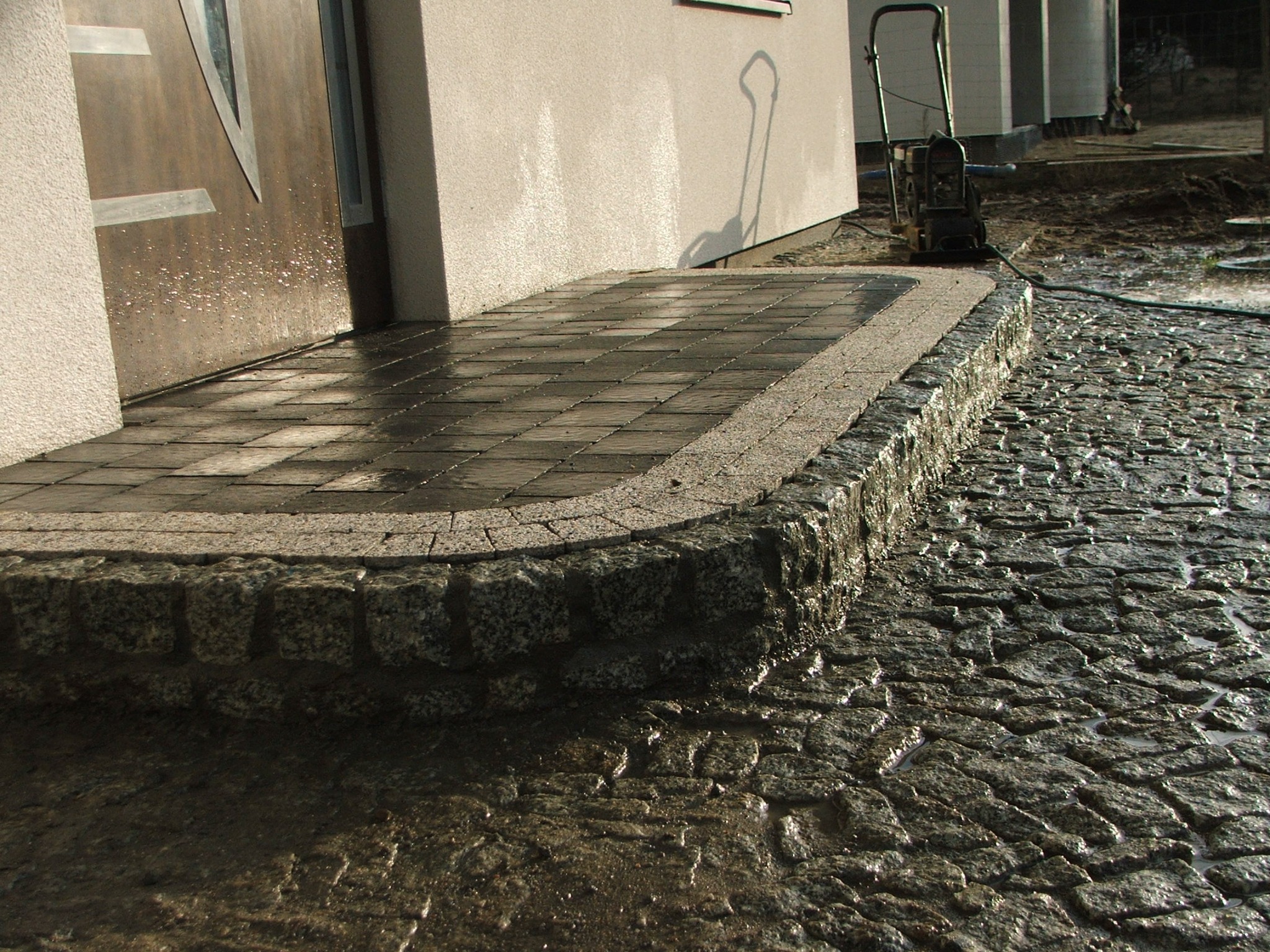 Ułożony chodnik z kostki brukowej przed wejściem do domu, z widoczną maszyną do zagęszczania gruntu w tle i mokrą nawierzchnią.