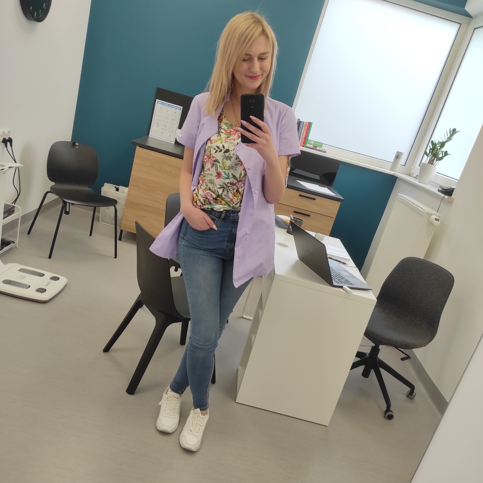 Blondynka w jeansach i fioletowym fartuchu medycznym robi selfie w gabinecie z wagą diagnostyczną, biurkiem z laptopem i oknem z rośliną w tle.